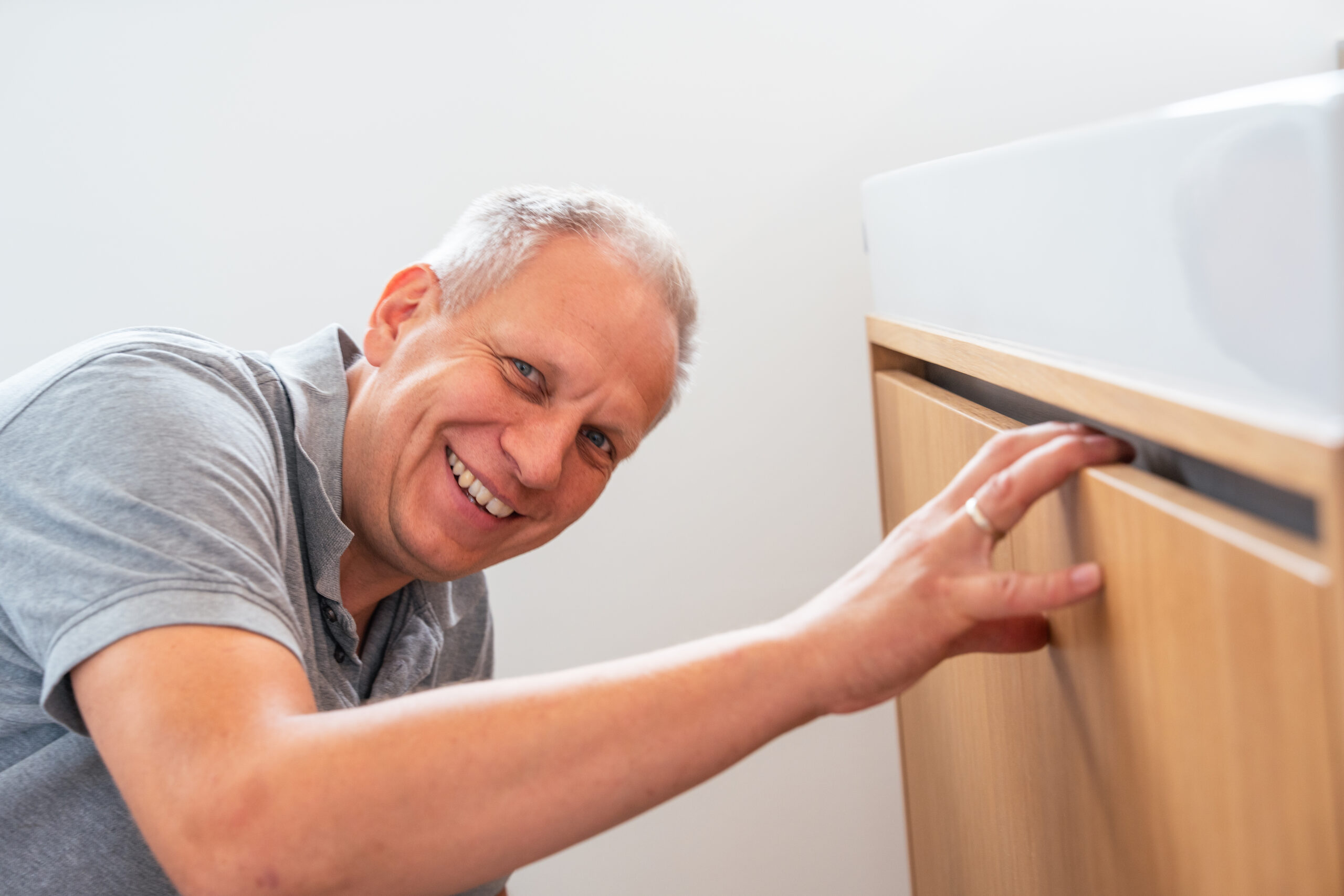 Tischlermeister lächelt in die Kamera, während er einen Holzschrank unter einem Waschbecken montiert.